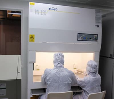 Two gowned technicians working side by side at a biosafety cabinet inside a cleanroom suite.