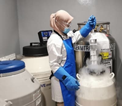 A gowned technician lifting a rack from an open liquid-nitrogen dewar, with vapour spilling over the rim.