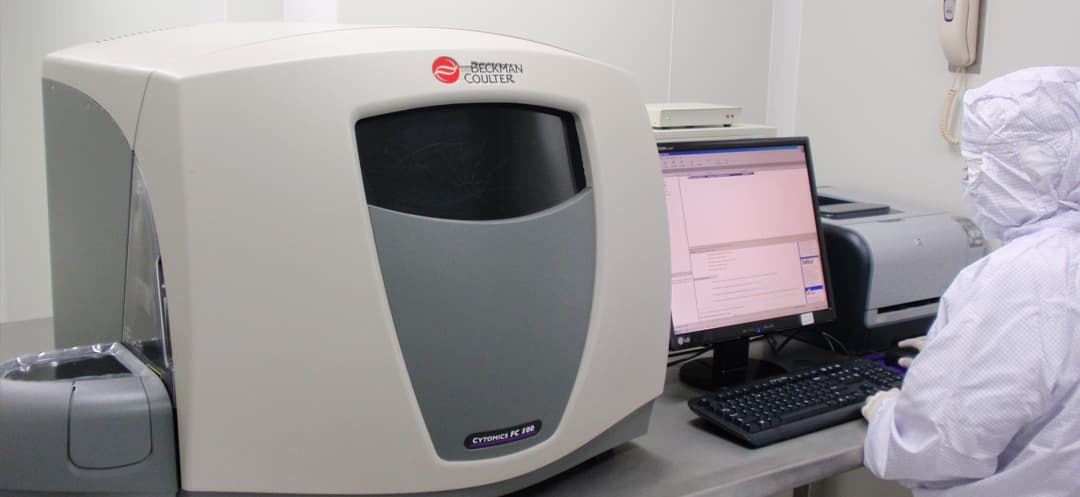 A gowned operator seated at a Beckman Coulter Cytomics FC 500 flow cytometer in the NiSCELL laboratory, reading the analysis on the workstation beside it.
