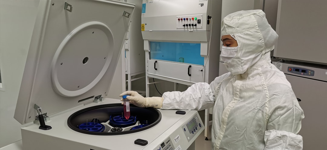 A gowned technician lowering a sample tube into an open floor-standing centrifuge inside a NiSCELL cleanroom, with a biosafety cabinet behind.