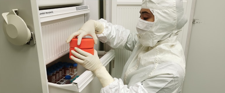 A gowned technician retrieving a labelled reagent box from a cleanroom freezer.